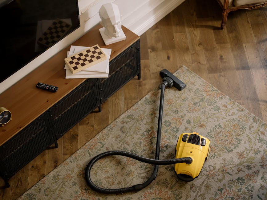 A woman, wearing a beige jacket and dark jeans, is engaged in surface cleaning with a vacuum cleaner in a living room. She is standing on a patterned area rug that covers a wooden floor. The vacuum cl