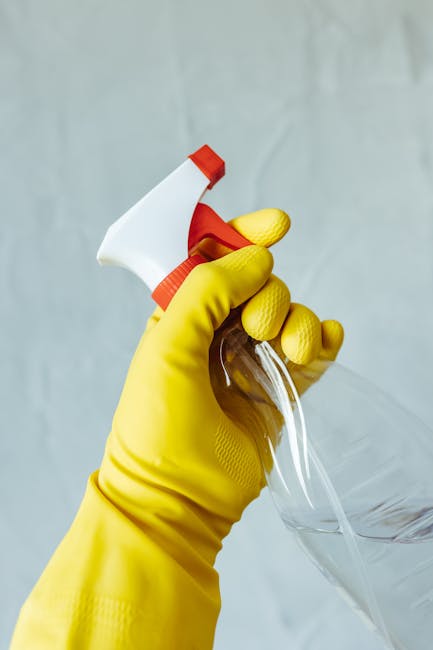 Close-up of a hand wearing a bright yellow cleaning glove gripping a white spray bottle with a red nozzle, aimed at a clear glass surface. The background is a neutral, slightly textured grey wall. The