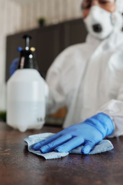 A person dressed in white protective clothing, including a face mask and blue gloves, is cleaning a wooden surface with a cloth in an indoor setting. Visible cleaning equipment such as a spray bottle 