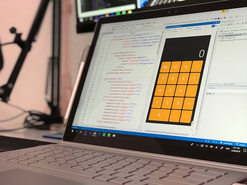 Close-up of a silver laptop computer displaying a coding editor on the left and a calculator app with orange buttons on the right, placed on a desk with a blurred background showing a tripod stand and