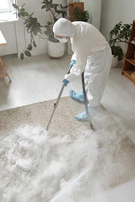 A person is vacuuming a patterned carpet in a room with natural light. The vacuum cleaner, with a black head and a long black tube, is positioned on the beige and muted green area rug, which features